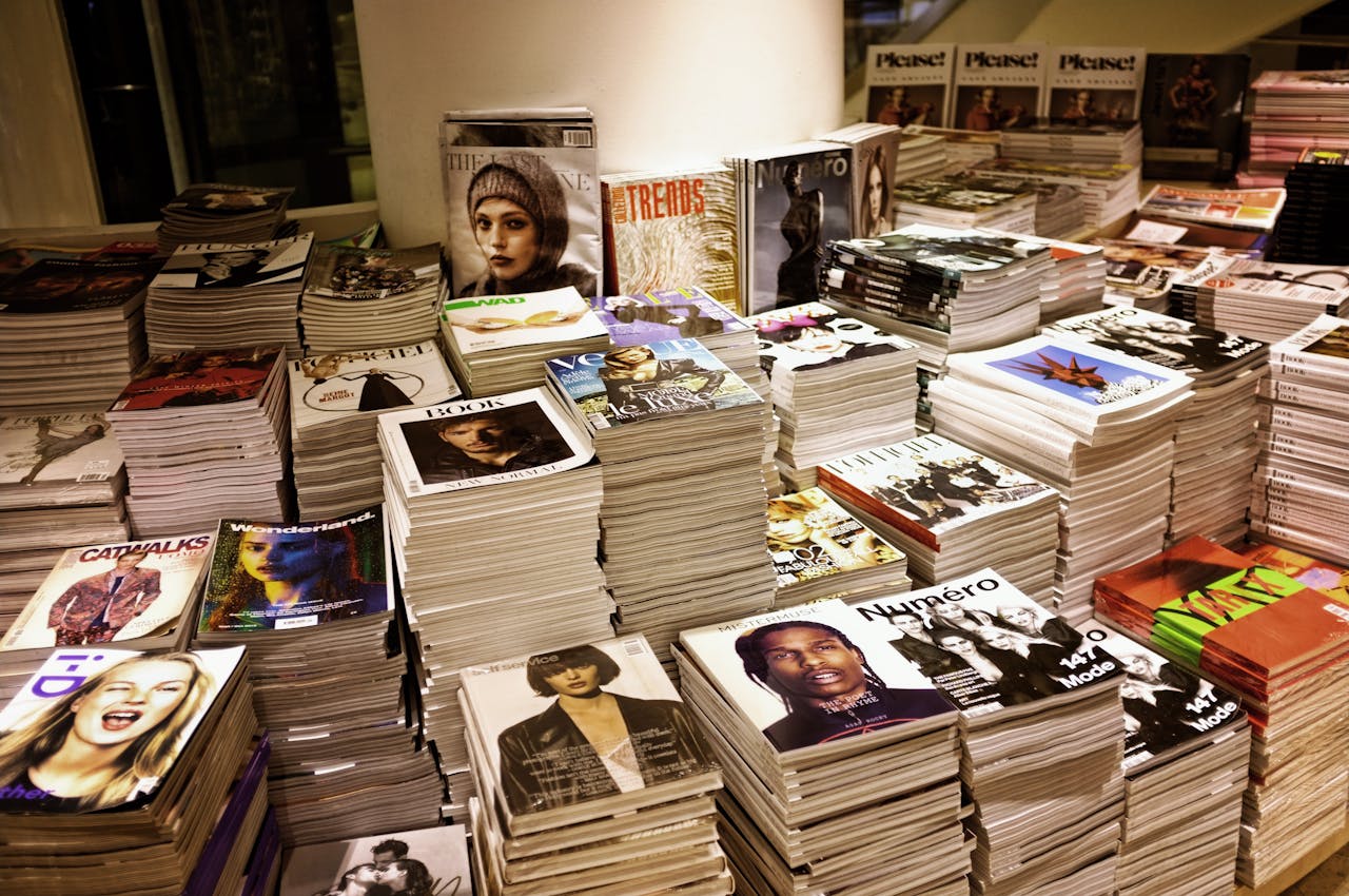 about-us Huge stacks of diverse fashion and lifestyle magazines piled high in an indoor store setting.